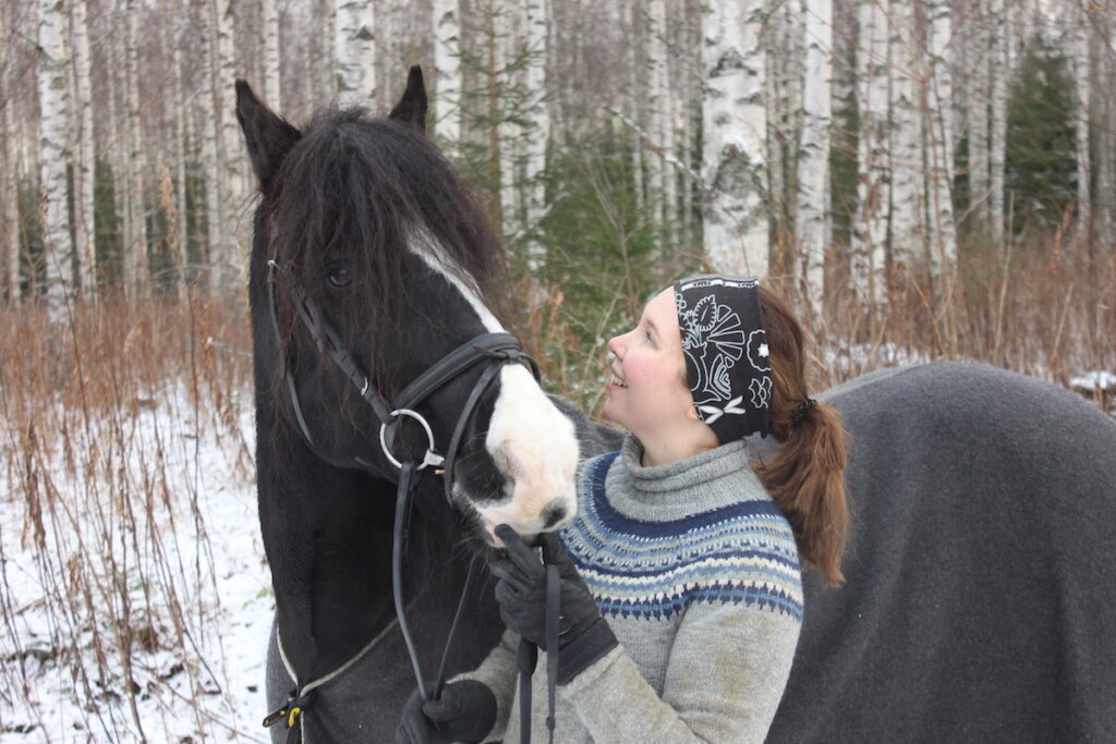 villapaitainen nuori nainen seisoo läsipää mustan irlannin cob hevosen kanssa lumisessa metsämaisemassa