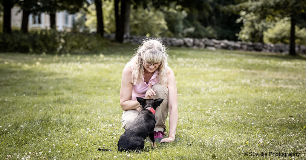 vaaleatukkainen nainen kyykyssä nurmikolla musta pieni koira edessään kesäisenä päivänä