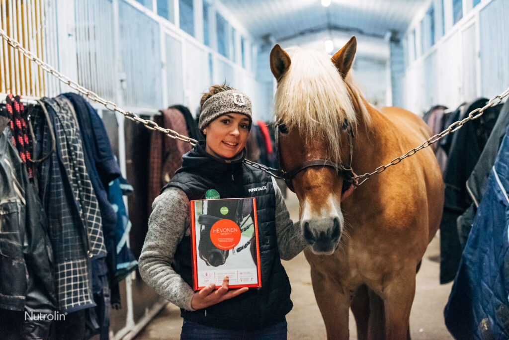 nuori nainen pitää sylissään Nutrolin Horse Sport -pakettia seistessään liinakkosuomenhevosorin vierellä tallin käytävällä