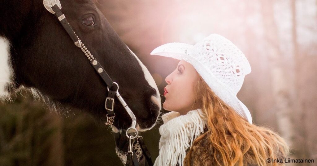 stetsonpäinen punatukkainen nainen suutelee mustavalkokirjavan hevosen turpaa